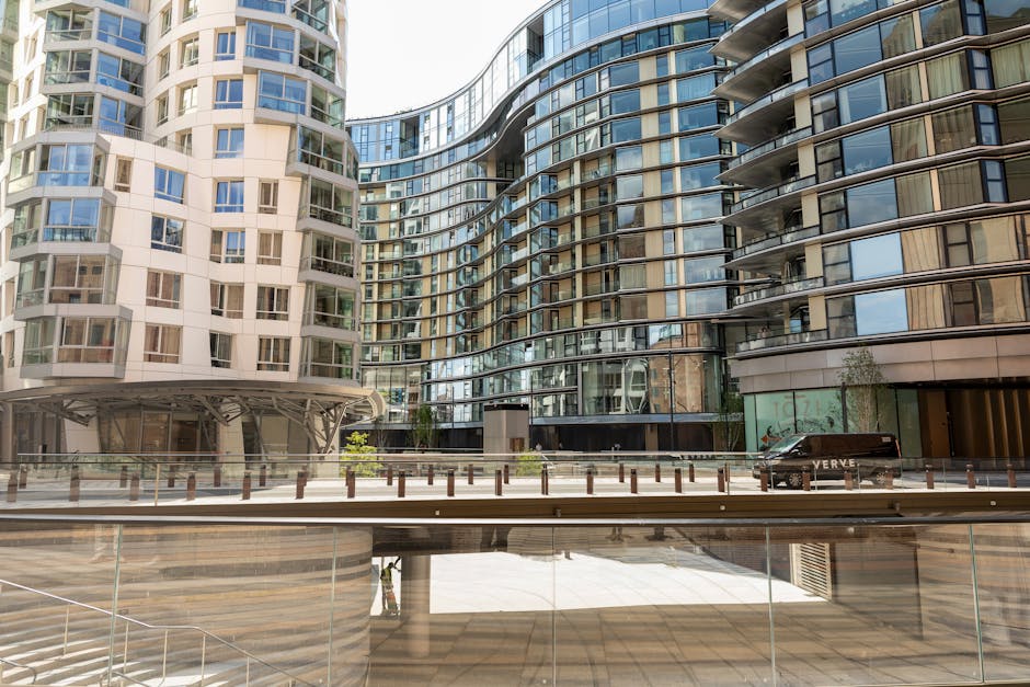 A modern multi-storey apartment building with curved glass facades and numerous balconies reflecting the surrounding environment. The building is situated in an urban area, with a paved street in front that has metal bollards and a black van parked along the curb. The scene is outdoors, during daylight, with ample natural light illuminating the building's contemporary architecture. The image may relate to house removals or relocation services provided by companies like manandvanelephantandcastle.org.uk, highlighting the context of moving or transporting furniture and personal belongings in a city setting.