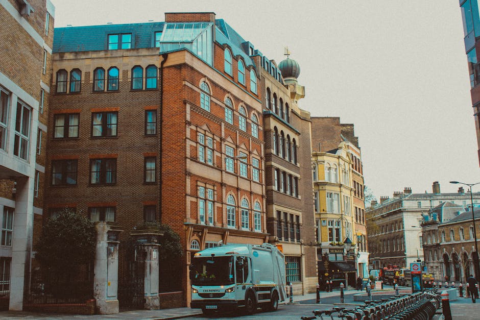 A white removal van with an open back section is parked on a city street in front of a row of multi-storey buildings, including a prominent red-brick structure with large windows and decorative arches
