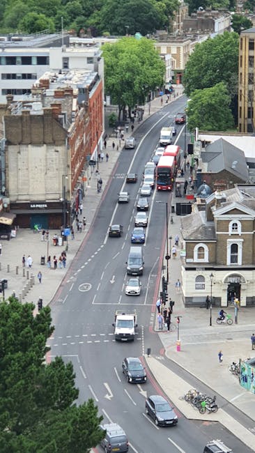 An aerial view of a busy urban street in Walworth, with several parked vehicles along the curb and moving traffic on the road, including cars and buses. The sidewalk features pedestrians walking, some carrying bags, and others waiting near a bus stop. There are various buildings lining the street, including a brick building on the left with signage, and a small historic-looking church or chapel with white walls and a pitched roof on the right. Mature trees with lush green foliage are visible along the street, providing some shade. The photograph appears to capture a moment during the daytime with natural lighting, highlighting activity related to house removals and the typical environment encountered during furniture transport and relocation logistics, with no movement of furniture or packing materials shown in the image. This scene exemplifies the urban setting where professional removals companies like [COMPANY_NAME] operate to facilitate home relocation and moving services within this area, supporting the logistics involved in packing, loading, and transportation to and from residential and commercial properties.