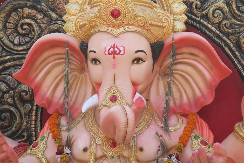 A close-up image of a colorful statue of Lord Ganesha, the Hindu deity with an elephant head, featuring a pink complexion and large, flared ears. The statue is adorned with intricate gold jewelry and decorative elements, including a red gemstone on the forehead and gold embellishments around the neck and head. Fresh flowers, such as marigolds and white blossoms, are draped around the statue's neck. The background shows a dark, ornate carved surface, highlighting the detailed craftsmanship of the idol. This image captures the spiritual and cultural significance of Ganesha, often associated with home prayers and religious ceremonies, which can be relevant when considering the importance of respectful handling during house removals or relocation services offered by [COMPANY_NAME], especially when moving religious artifacts or decorative statues.