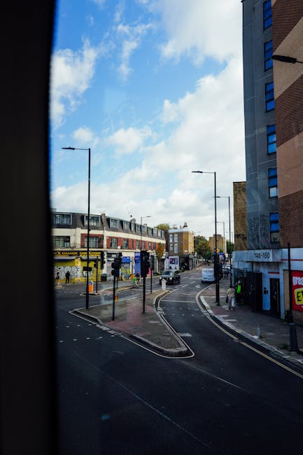 An aerial view of a busy urban street in Walworth, with several parked vehicles along the curb and moving traffic on the road, including cars and buses. The sidewalk features pedestrians walking, some carrying bags, and others waiting near a bus stop. There are various buildings lining the street, including a brick building on the left with signage, and a small historic-looking church or chapel with white walls and a pitched roof on the right. Mature trees with lush green foliage are visible along the street, providing some shade. The photograph appears to capture a moment during the daytime with natural lighting, highlighting activity related to house removals and the typical environment encountered during furniture transport and relocation logistics, with no movement of furniture or packing materials shown in the image. This scene exemplifies the urban setting where professional removals companies like [COMPANY_NAME] operate to facilitate home relocation and moving services within this area, supporting the logistics involved in packing, loading, and transportation to and from residential and commercial properties.